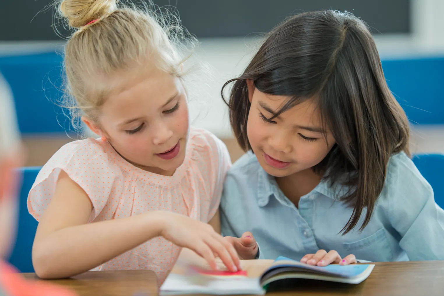 Two young students sit at a desk and look down and point at a book.