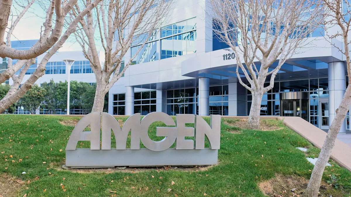 The exterior of Amgen's office building in South San Francisco, with a sculpted company sign in the foreground and leafless trees on a grassy lawn.