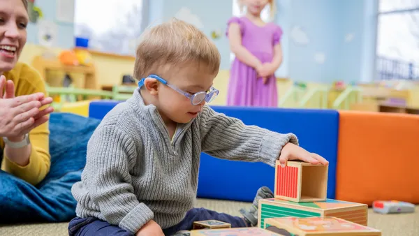 A baby is sitting on the floor playing with blocks. An adult is watching and clapping and a young student is standing watching in the background.