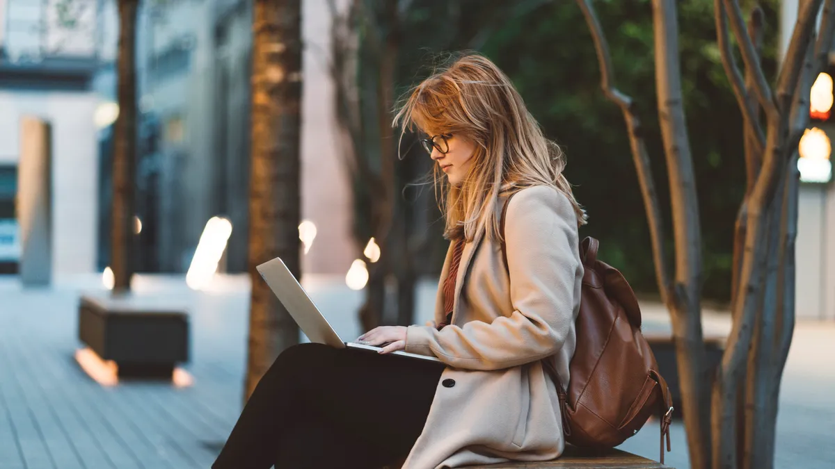 A young woman works on her computer on a bench.