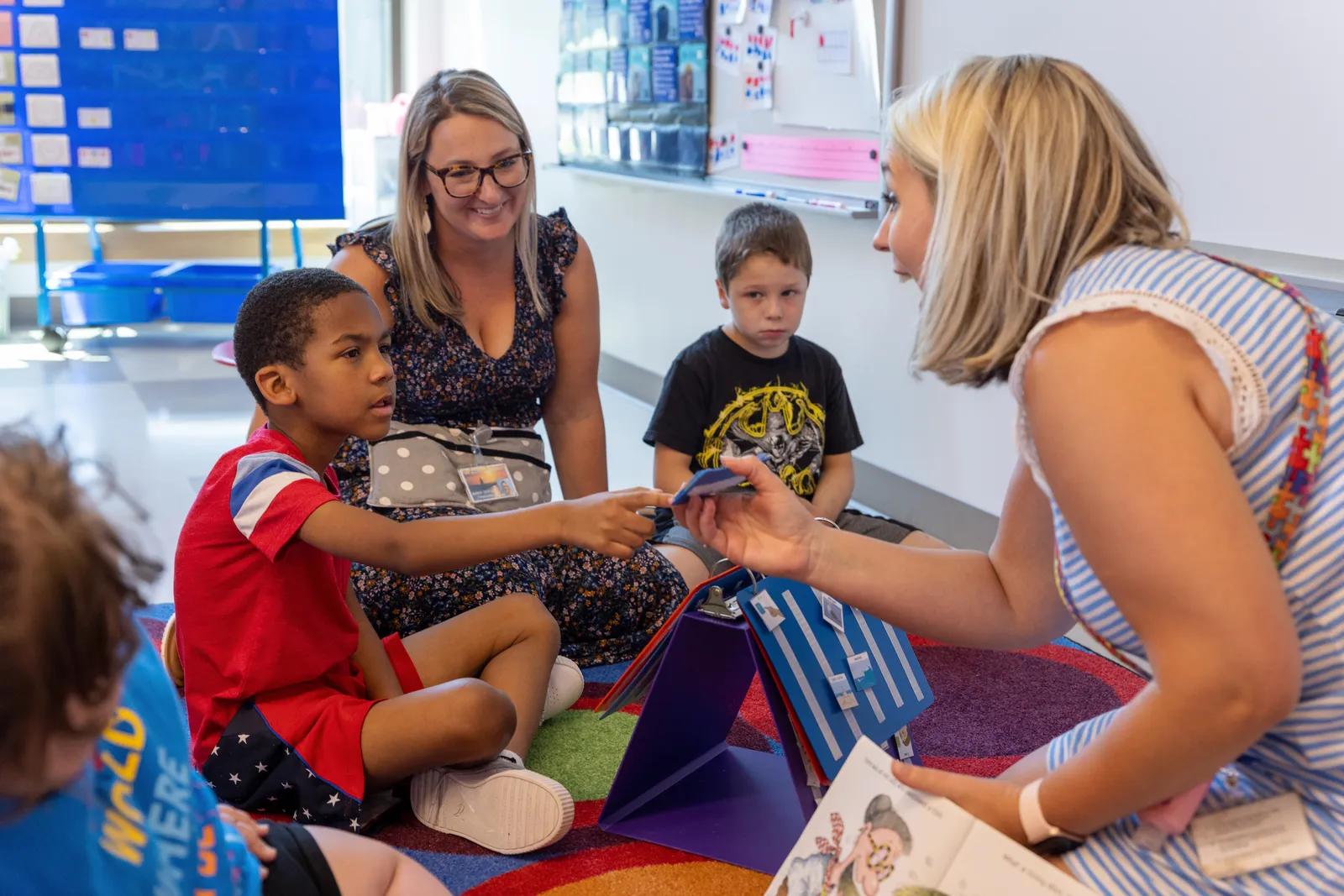 two young students are sitting on the floor in a classroom. Two adults are sitting with them and one student is pointing to a book being held by one of the adults.