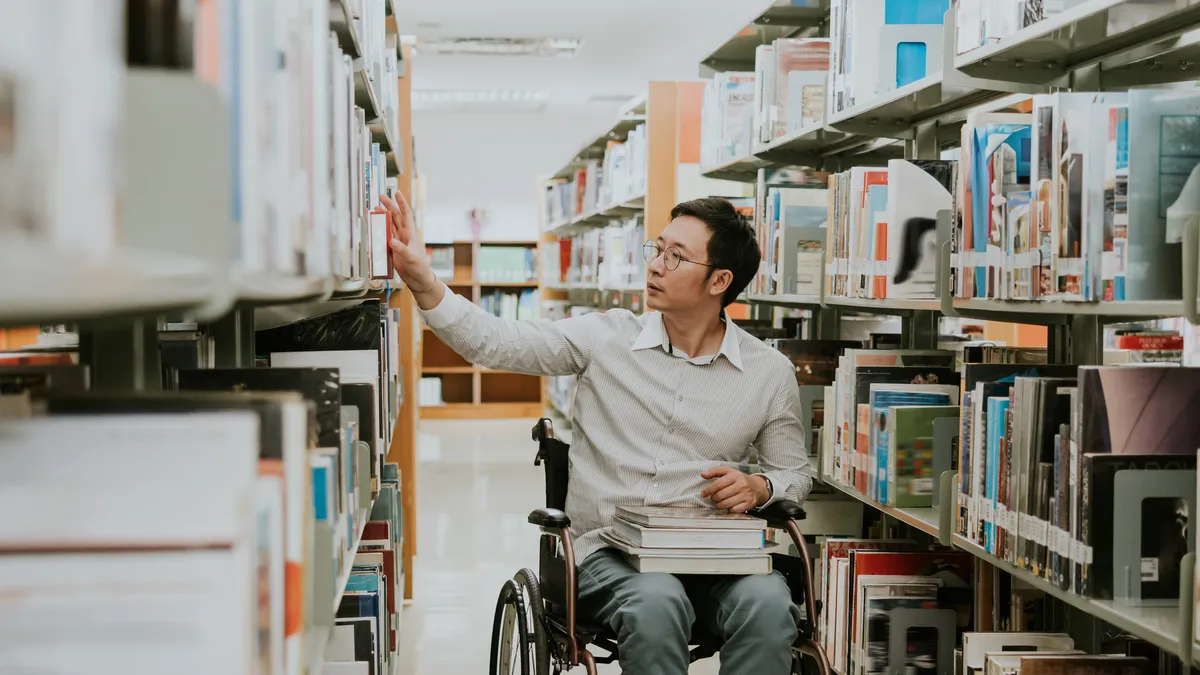 An Asian person in a wheelchair wheels through a library