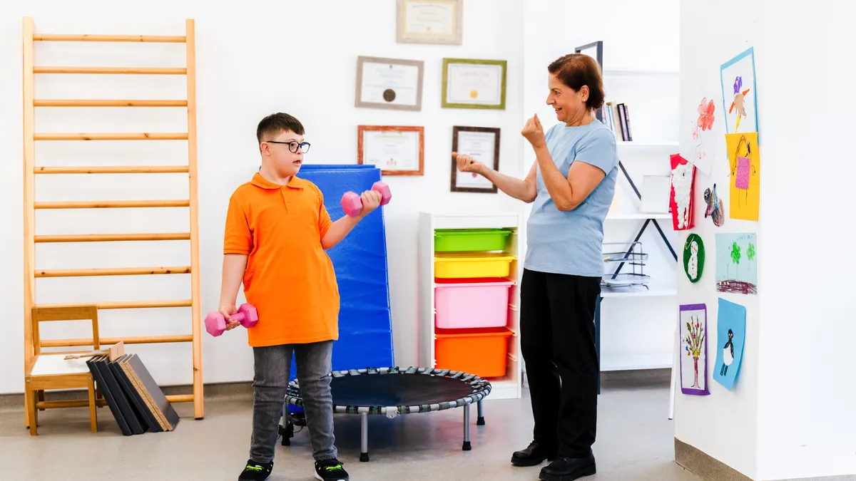 a students is standing in a room with exercise equipment and hold weights in each arm. An adult stands nearby demonstrating how to life the weights