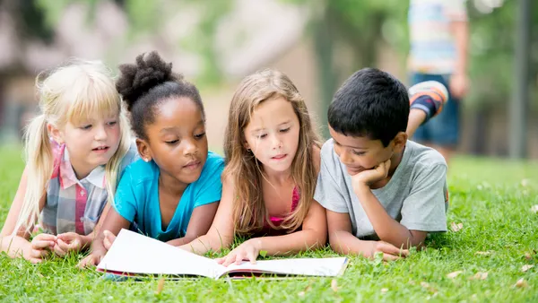 Four students are laying on their stomachs outside on the grass. They are looking at a book in front of them.