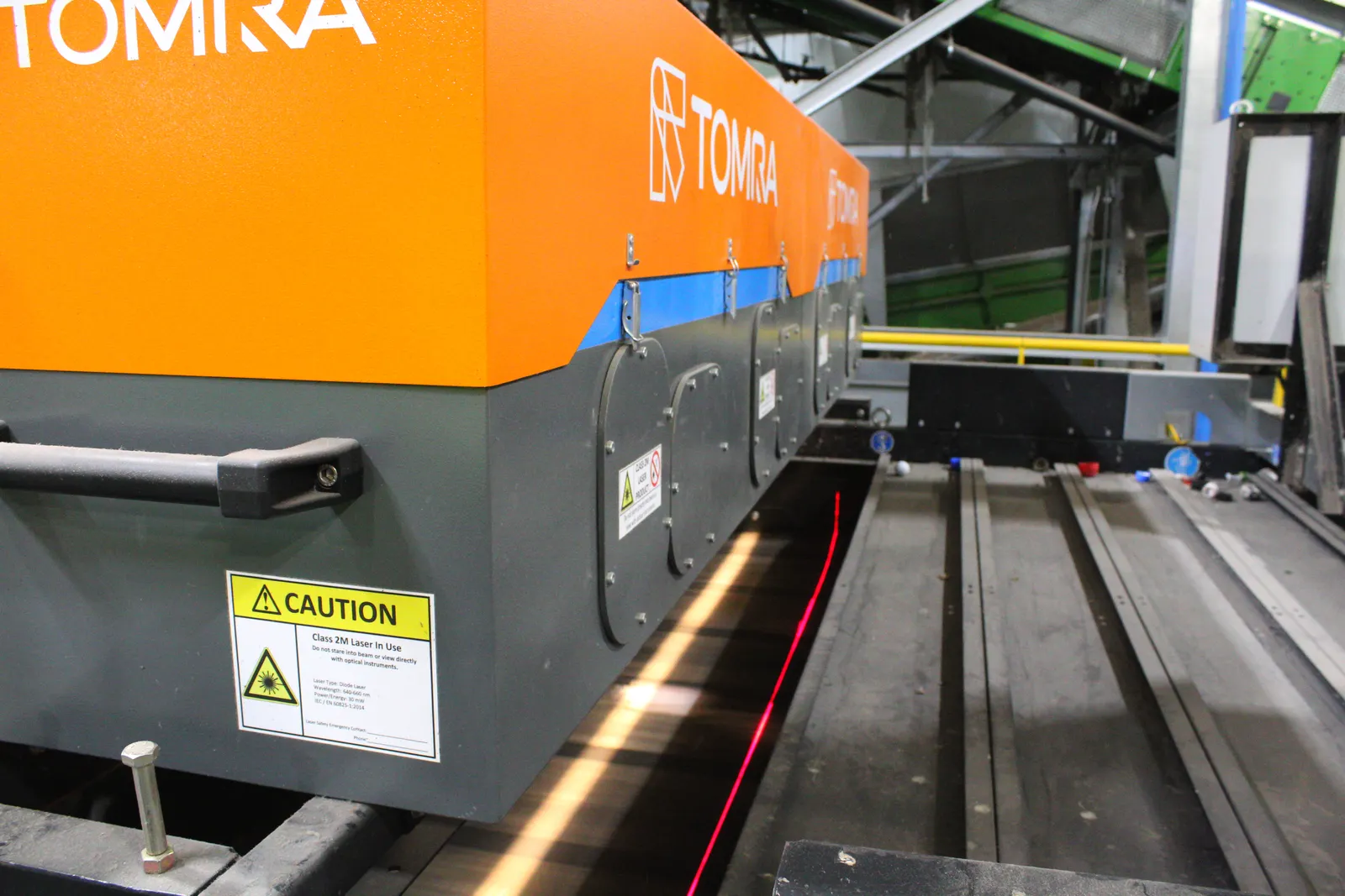 A piece of industrial equipment with Tomra's brand name shines a white light and laser onto a conveyor belt underneath.