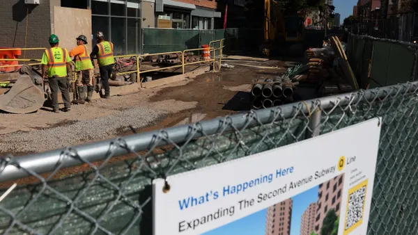 A fence in the reads"What's Happening Here: The Second Avenue Subway" with three construction workers standing on dirt next to a small brick building.