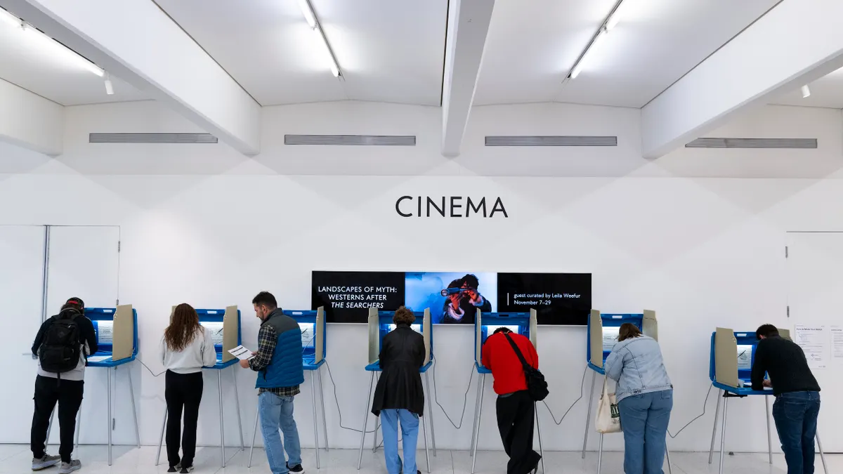 People stand at voting booths in a large white room. The word "Cinema" is on the wall above them.