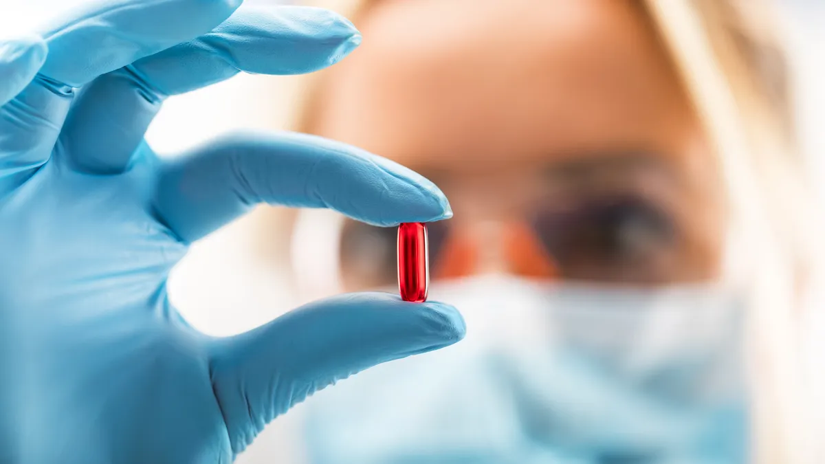 A scientist with protective eyeglasses and mask holding a red transparent pill with fingers in gloves in the pharmaceutical research laboratory