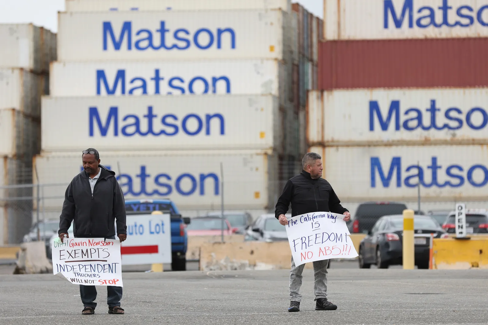 Two people hold signs that read, "the california way is freedom no AB5!!!" in front of shipping containers.