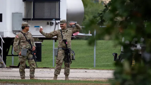 Two people in camouflage uniforms. One is taking a selfie with a smartphone.