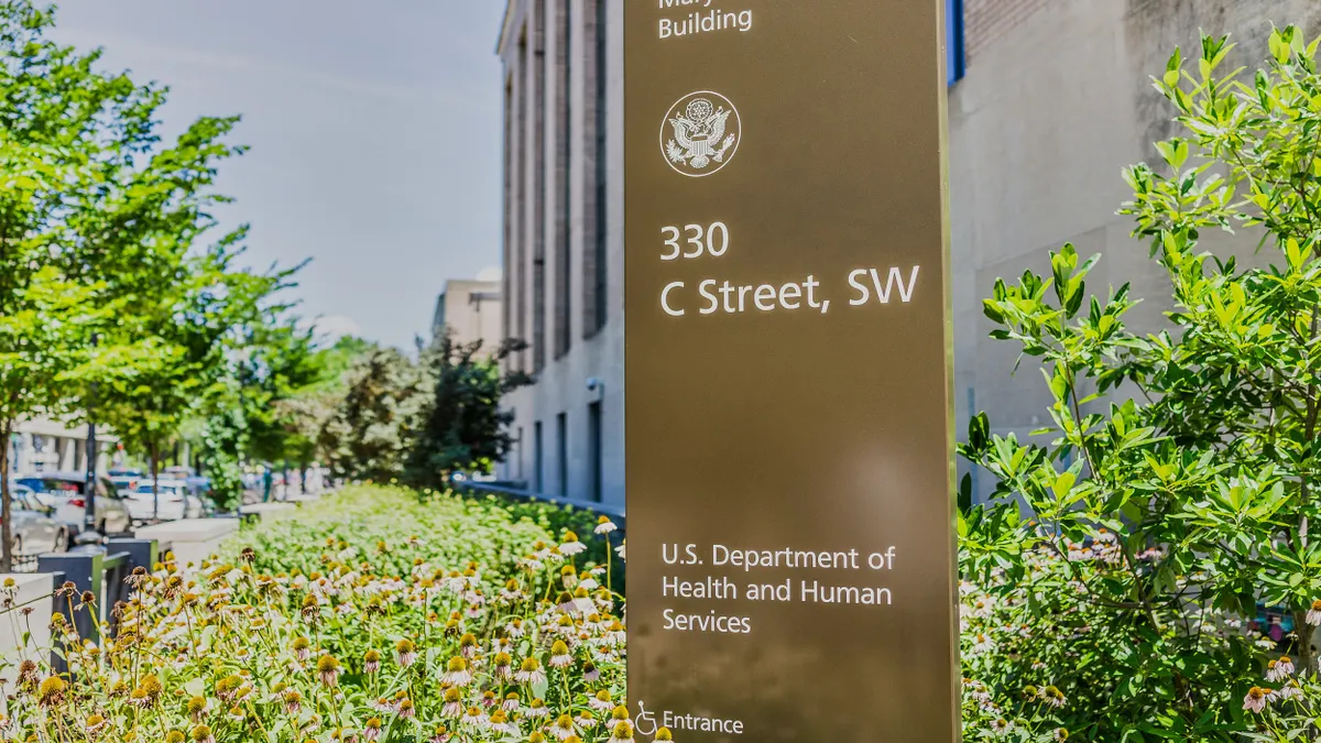A metal freestanding sign that reads, "Mary E Switzer Building, 330 C Street, SW, U.S. Department of Health and Human Services, Entrance" is surrounded by a brush with a building in the background. 