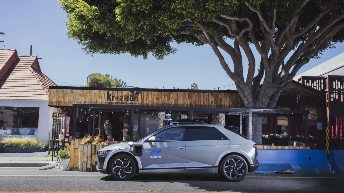 A gray four-door SUV marked as a Motional driverless vehicles is parked in front of a wood-sheathed restaurant with outdoor seating.