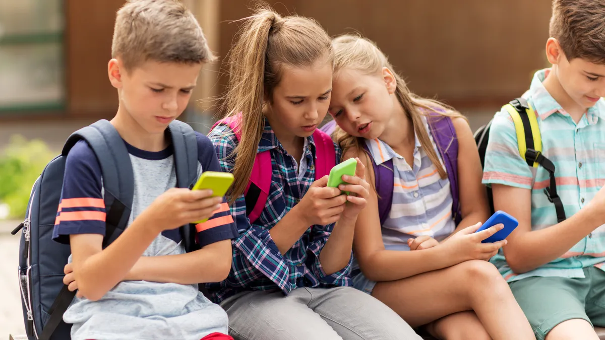 Four students with backpacks sit outside of a school looking at their cellphones.