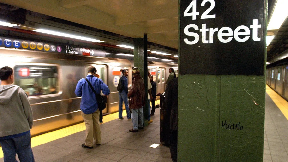 People stand at a subway station identified as 42nd Street in New York City by a large sign on a concrete pillar. A silver train is  arriving or departing.