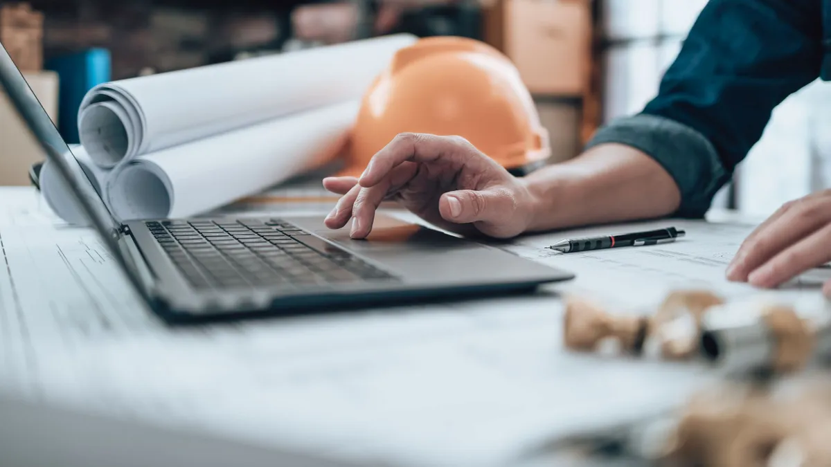 Engineer works at a table with a laptop, papers and a hard hat.