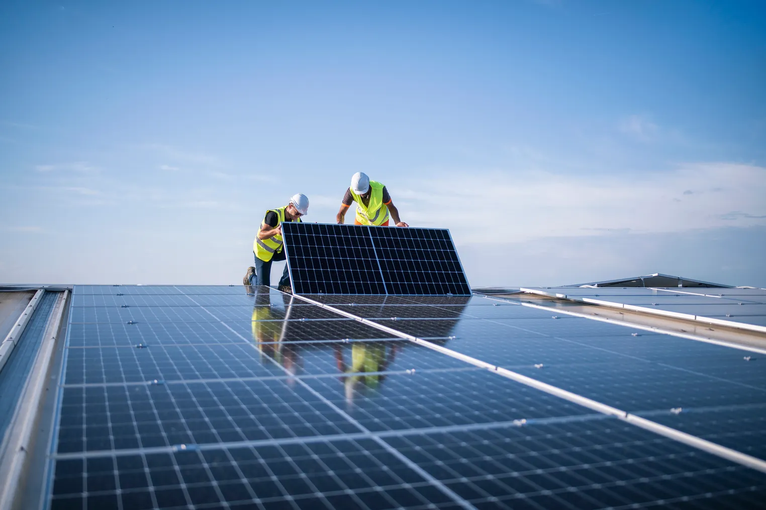 Two workers in hardhats and high-vis vests install a solar panel on a roof.