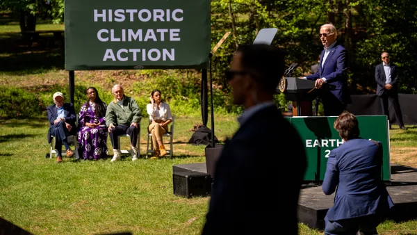 Four people, including members of Congress, sit in chairs under a sign that reads "President Joe Biden Historic Climate Action." Biden stands on a small stage to the right.