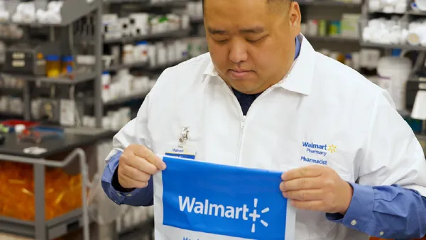 A pharmacist holds a bag, with shelves of medications in the background.