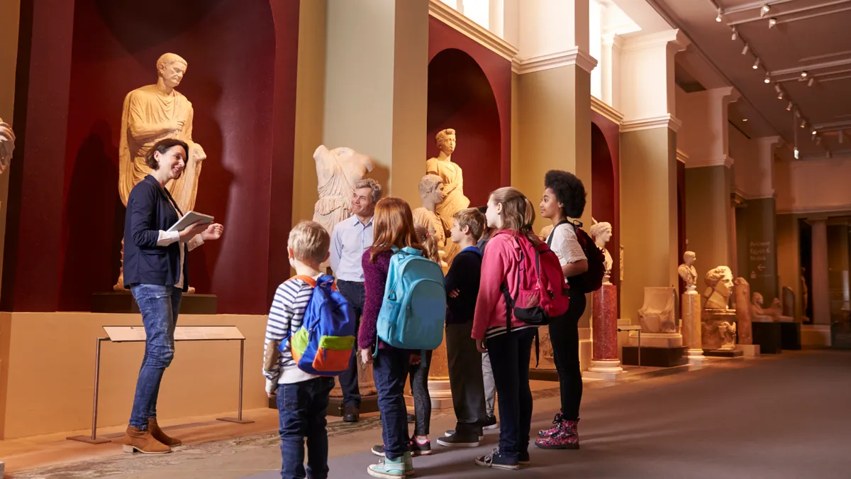 A group of students are shown on a trip to a museum in which a tour guide is explaining Greco-Roman statues to them.