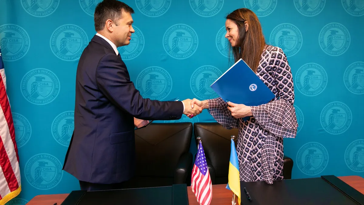 A man and a woman shake hands in front of a desk that has flags from the U.S. and Ukraine. The people are in front of a blue background with CISA logos.